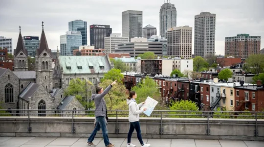 Deux personnes observent depuis un point de vue élevé le panorama de Montréal où se succèdent vieux quartiers, tours du centre-ville et maisons résidentielles à escaliers extérieurs.
