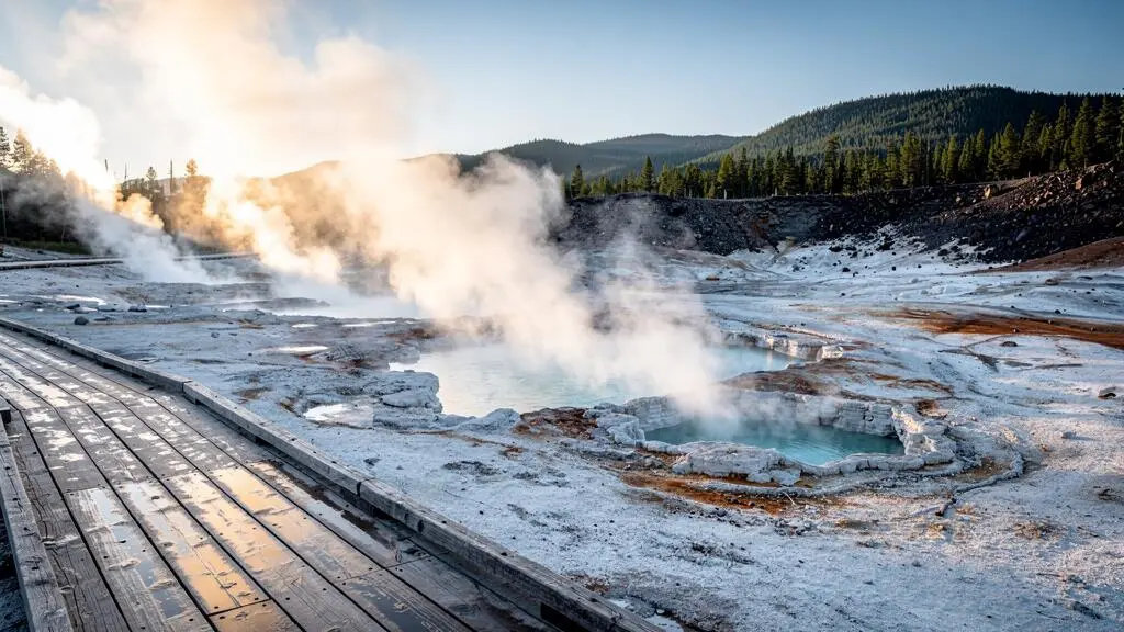 Un bassin géothermique volcanique au petit matin avec des geysers qui expulsent des colonnes de vapeur sous une lumière dorée.