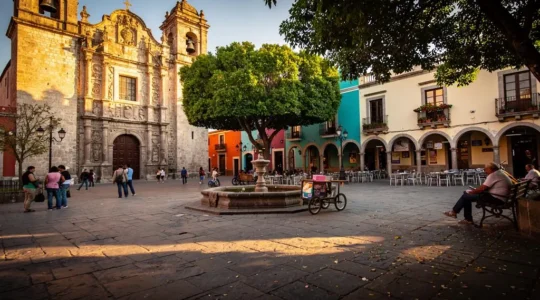 Place principale d'une ville coloniale mexicaine au coucher du soleil, avec la cathédrale, les arcades commerçantes et des passants traversant la place