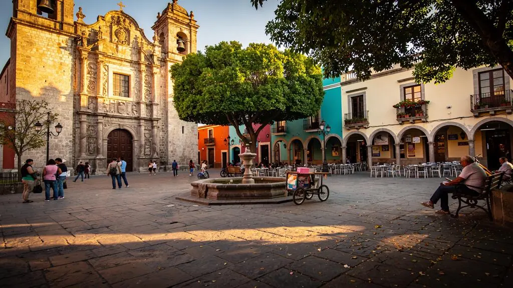 Place principale d'une ville coloniale mexicaine au coucher du soleil, avec la cathédrale, les arcades commerçantes et des passants traversant la place