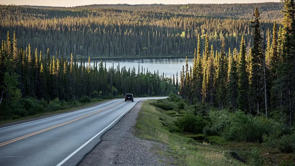 Une route traverse une immense forêt boréale d'épinettes canadiennes avec un lac glaciaire et un véhicule en trajet routier.