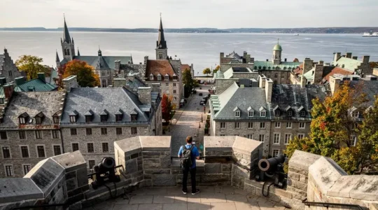 Un voyageur observe depuis les remparts en pierre les toits et rues anciennes d'une ville historique québécoise bordée par le fleuve.